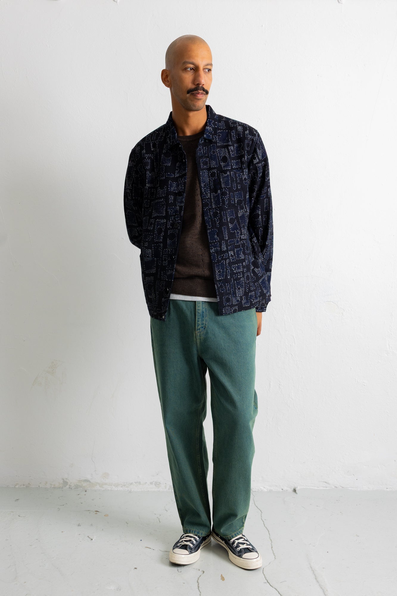 A man stands against a white wall wearing a dark navy patterned overshirt over a dark brown t-shirt, green pants, and black and white sneakers.