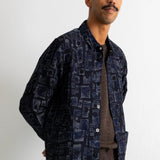 A man wearing a dark navy blue patterned button-up shirt jacket over a brown sweater and teal jeans, standing against a plain white wall.