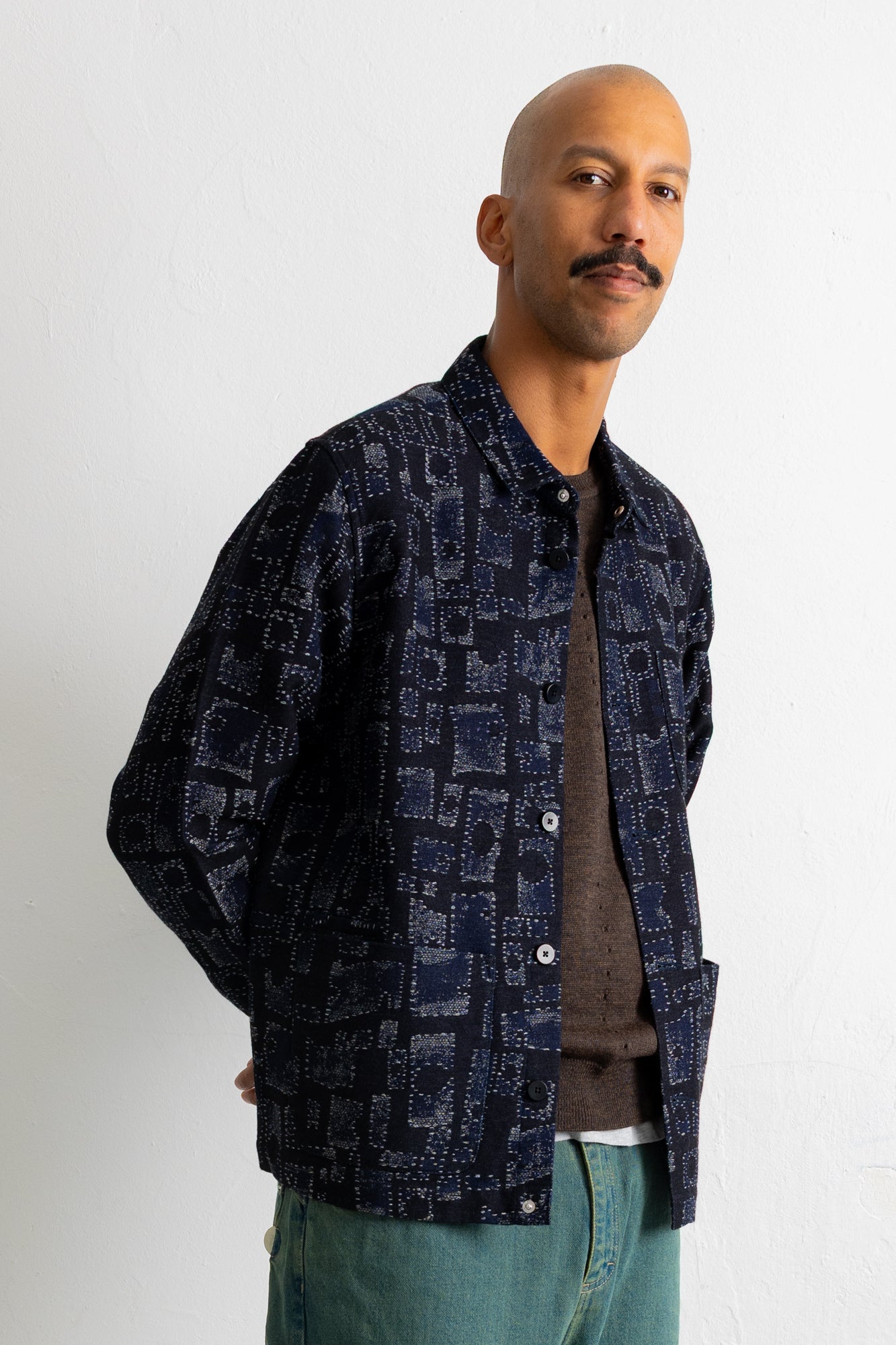 A man wearing a dark navy blue patterned button-up shirt jacket over a brown sweater and teal jeans, standing against a plain white wall.