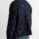 A man shown from the back wearing a dark navy blue patterned shirt jacket and teal pants, photographed against a plain white wall.