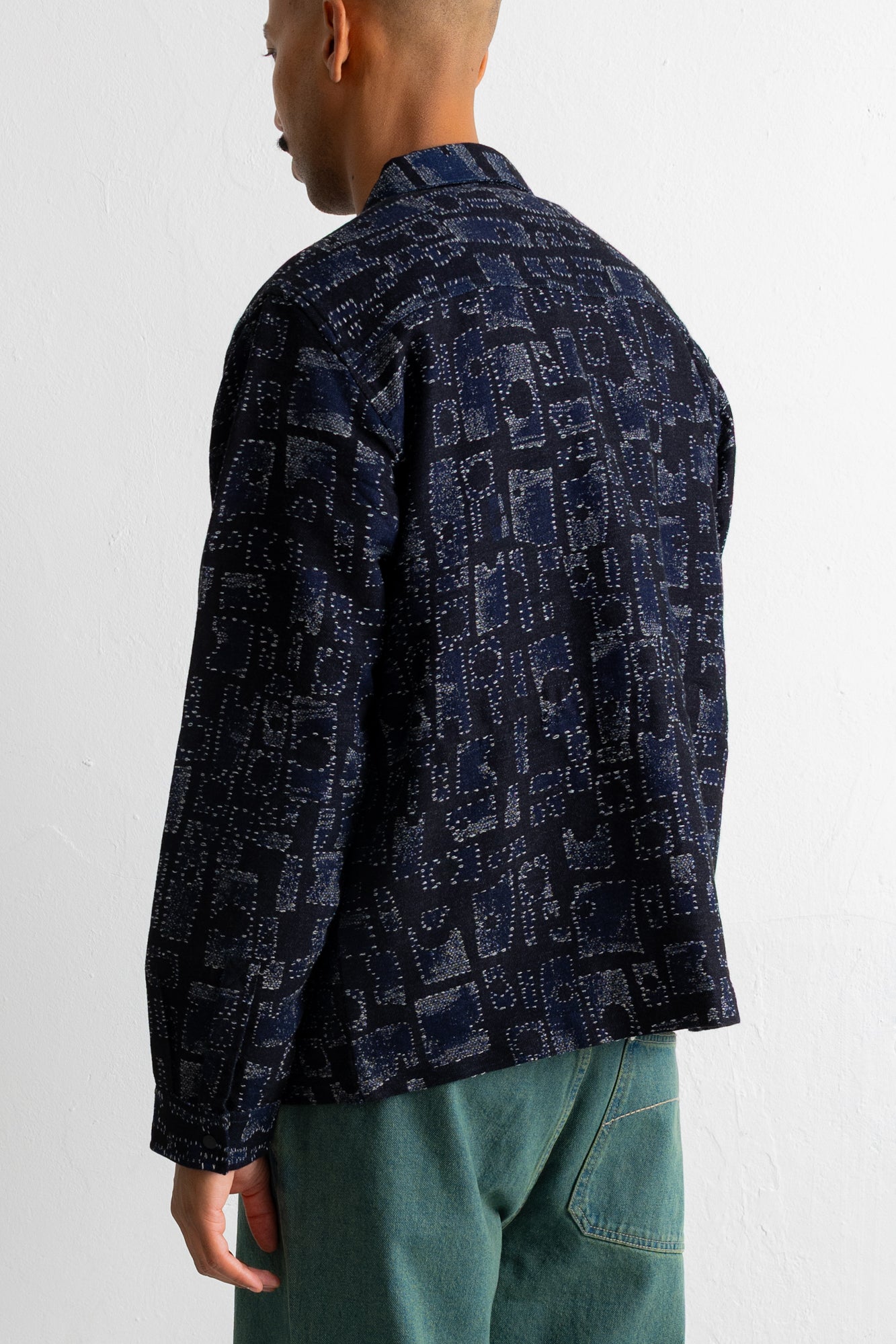 A man shown from the back wearing a dark navy blue patterned shirt jacket and teal pants, photographed against a plain white wall.