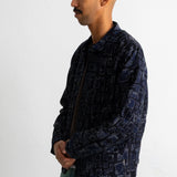 A man in profile wearing a dark navy blue patterned jacket and teal pants, standing against a plain white wall.