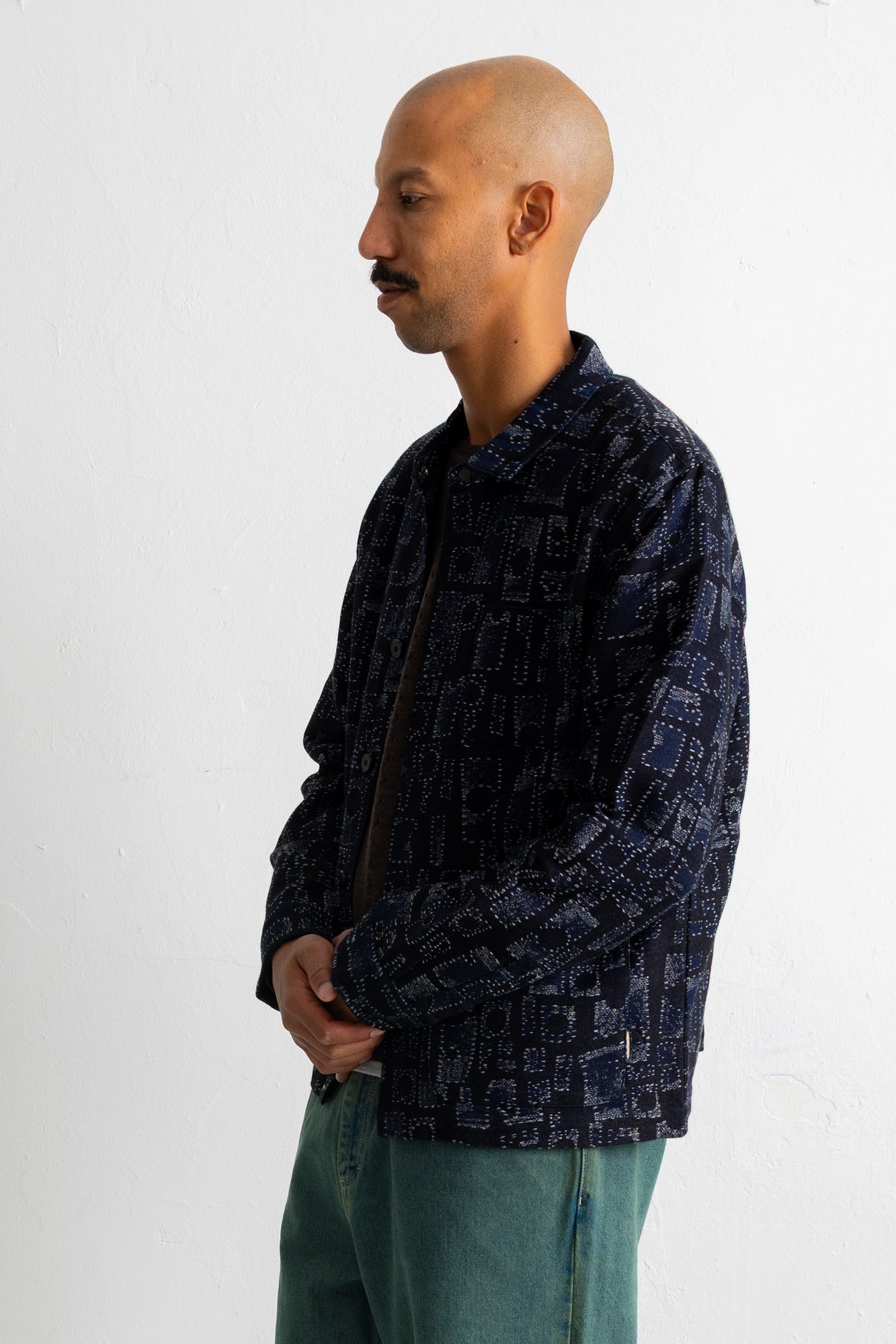 A man in profile wearing a dark navy blue patterned jacket and teal pants, standing against a plain white wall.