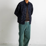 A man wearing a dark navy patterned button-up shirt jacket over a brown t-shirt, paired with green pants and black-and-white sneakers, standing against a white wall.
