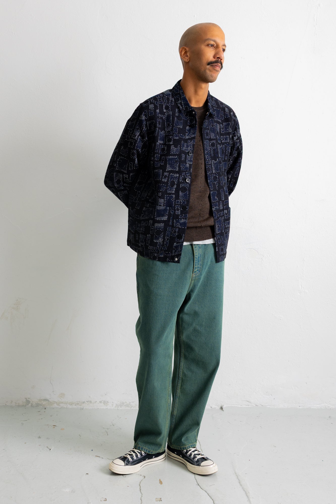 A man wearing a dark navy patterned button-up shirt jacket over a brown t-shirt, paired with green pants and black-and-white sneakers, standing against a white wall.