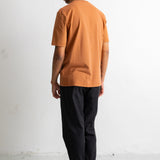 A man standing with his back to the camera, wearing a muted orange short-sleeve t-shirt, black pants, and black low-top sneakers with white soles, set against a plain white studio background.