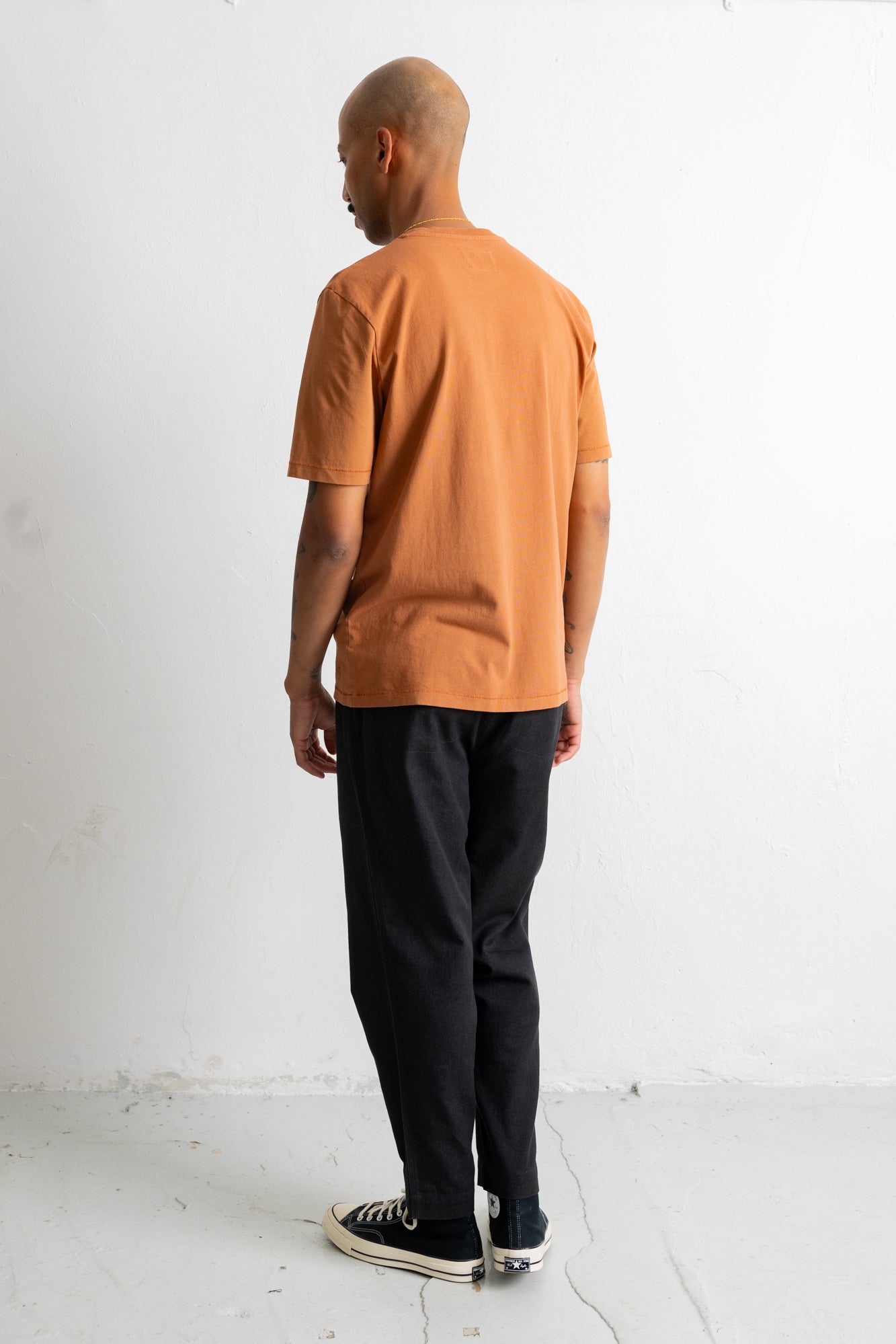 A man standing with his back to the camera, wearing a muted orange short-sleeve t-shirt, black pants, and black low-top sneakers with white soles, set against a plain white studio background.