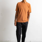 A man wearing a short-sleeve crewneck t-shirt in a muted orange, paired with black trousers and black-and-white sneakers, standing against a white wall.