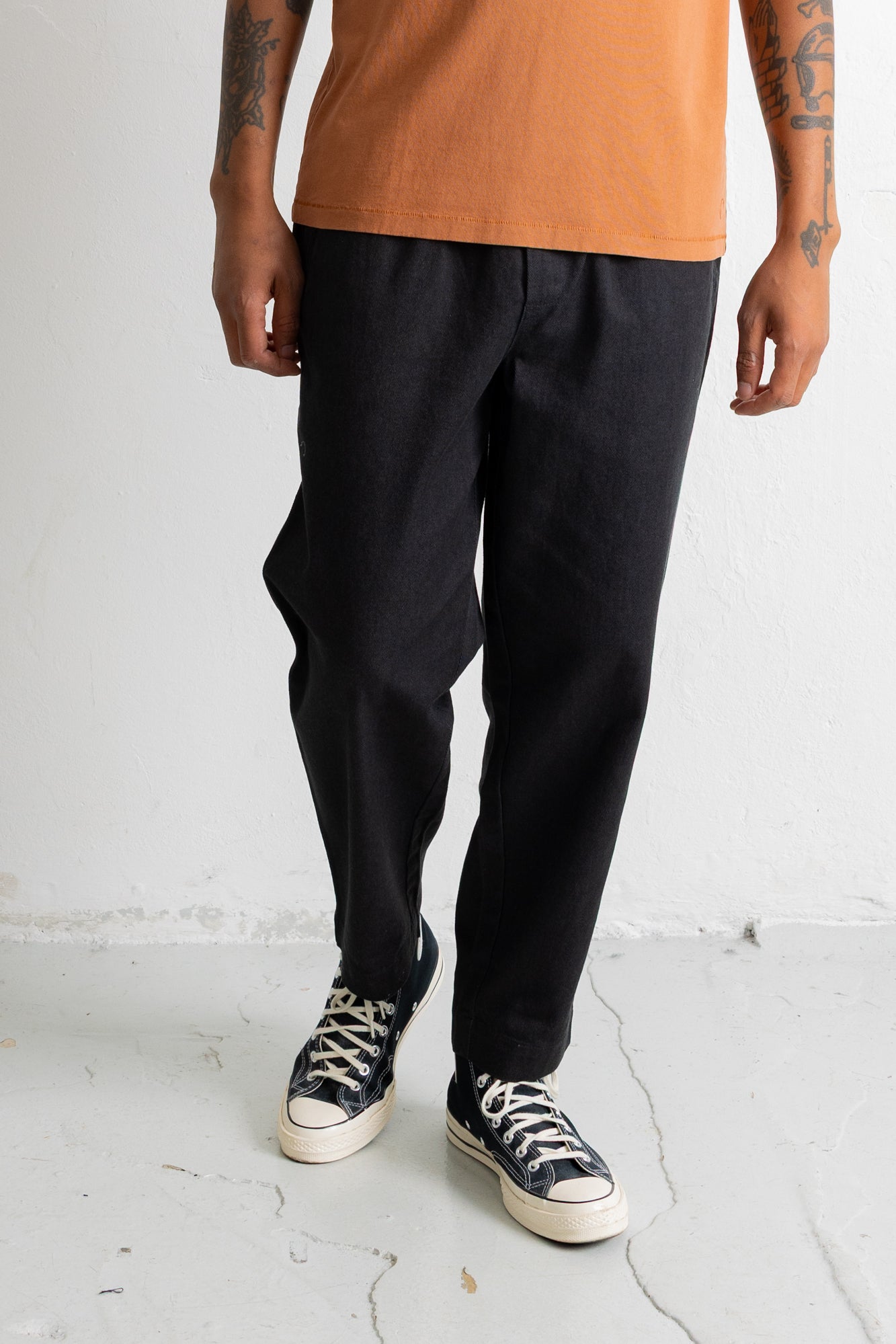 A person wearing loose black pants with an orange t-shirt and black low-top sneakers, standing against a white wall.