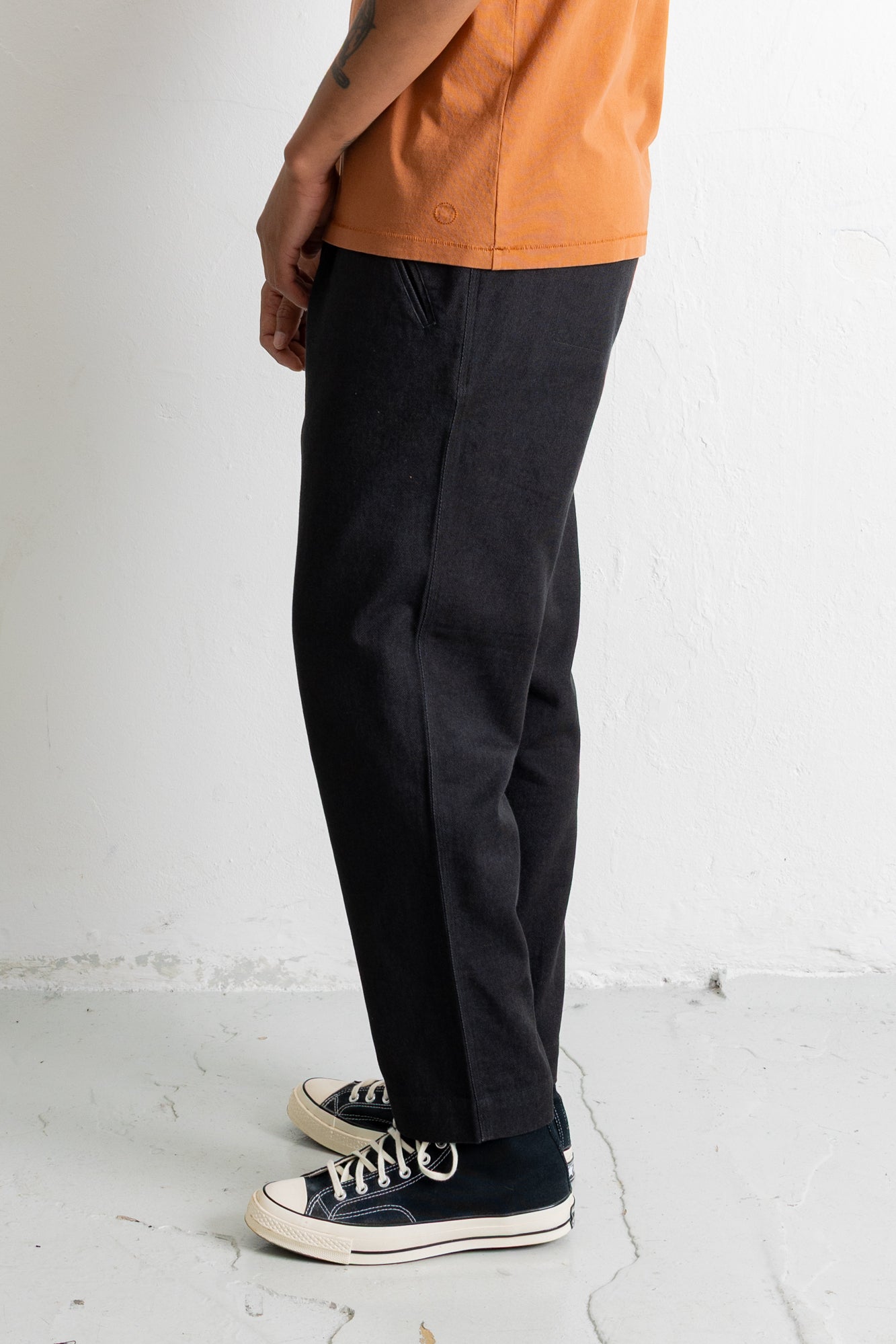 A person shown from the waist down wearing dark charcoal gray relaxed-fit pants, an orange t-shirt, and black canvas sneakers with white laces, standing against a white wall.