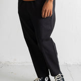 A person wearing loose black pants, a rust-orange t-shirt, and black Converse high-top sneakers, standing against a white textured wall.