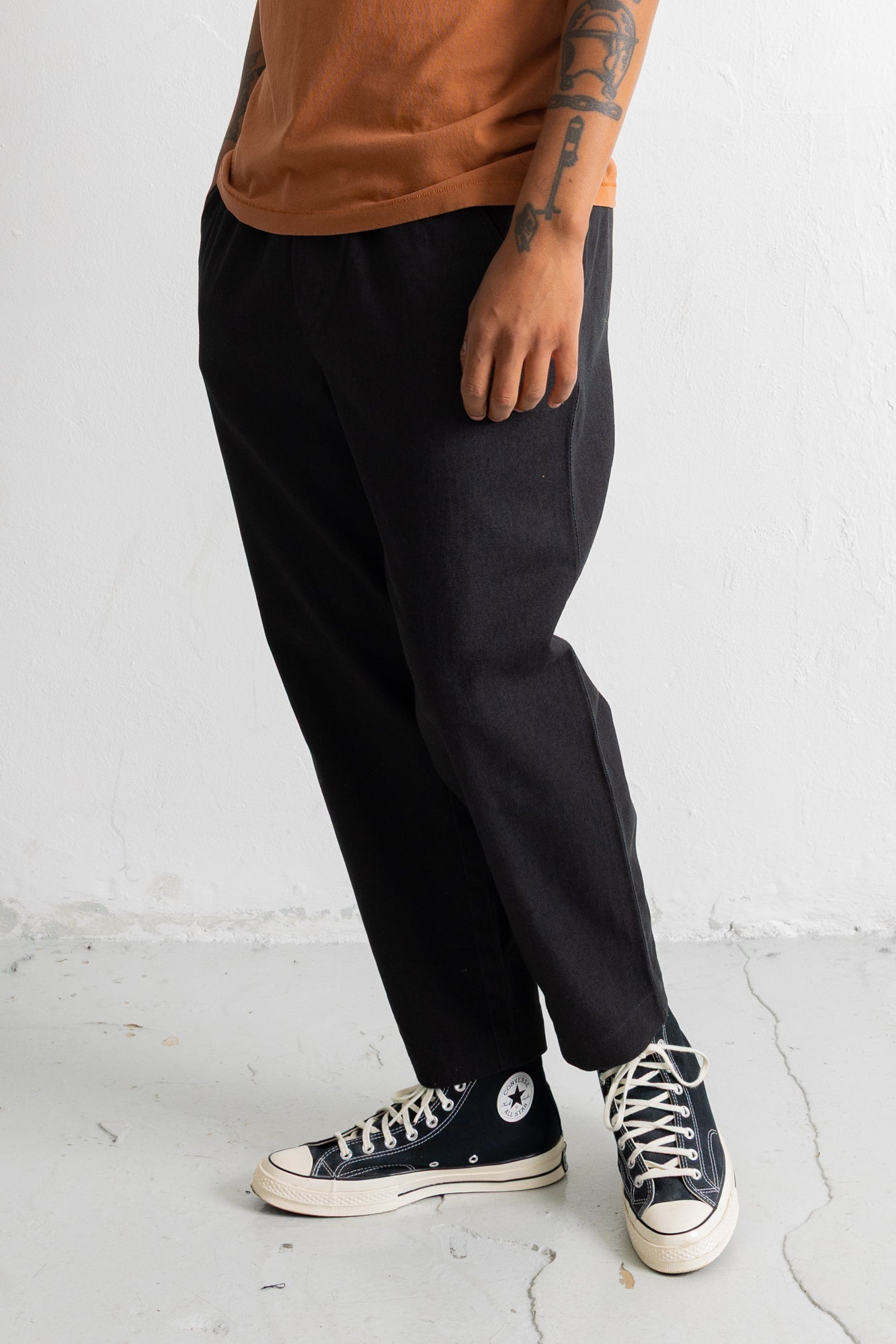A person wearing loose black pants, a rust-orange t-shirt, and black Converse high-top sneakers, standing against a white textured wall.