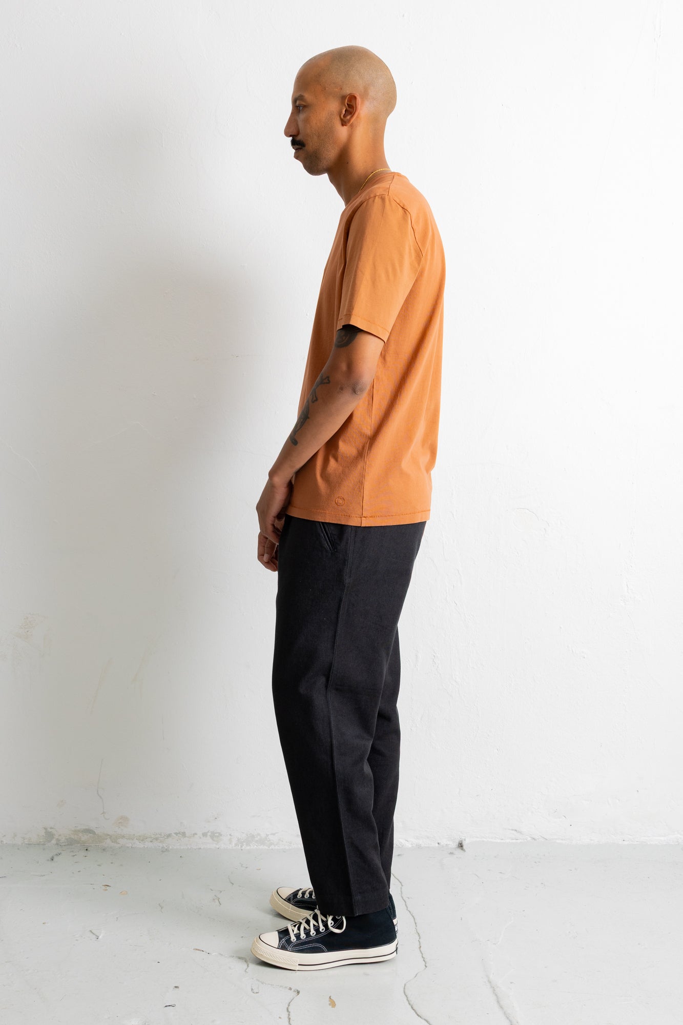 A man in profile wearing a short-sleeve crewneck t-shirt in a muted orange color, paired with dark charcoal pants and black canvas sneakers, standing against a white wall.