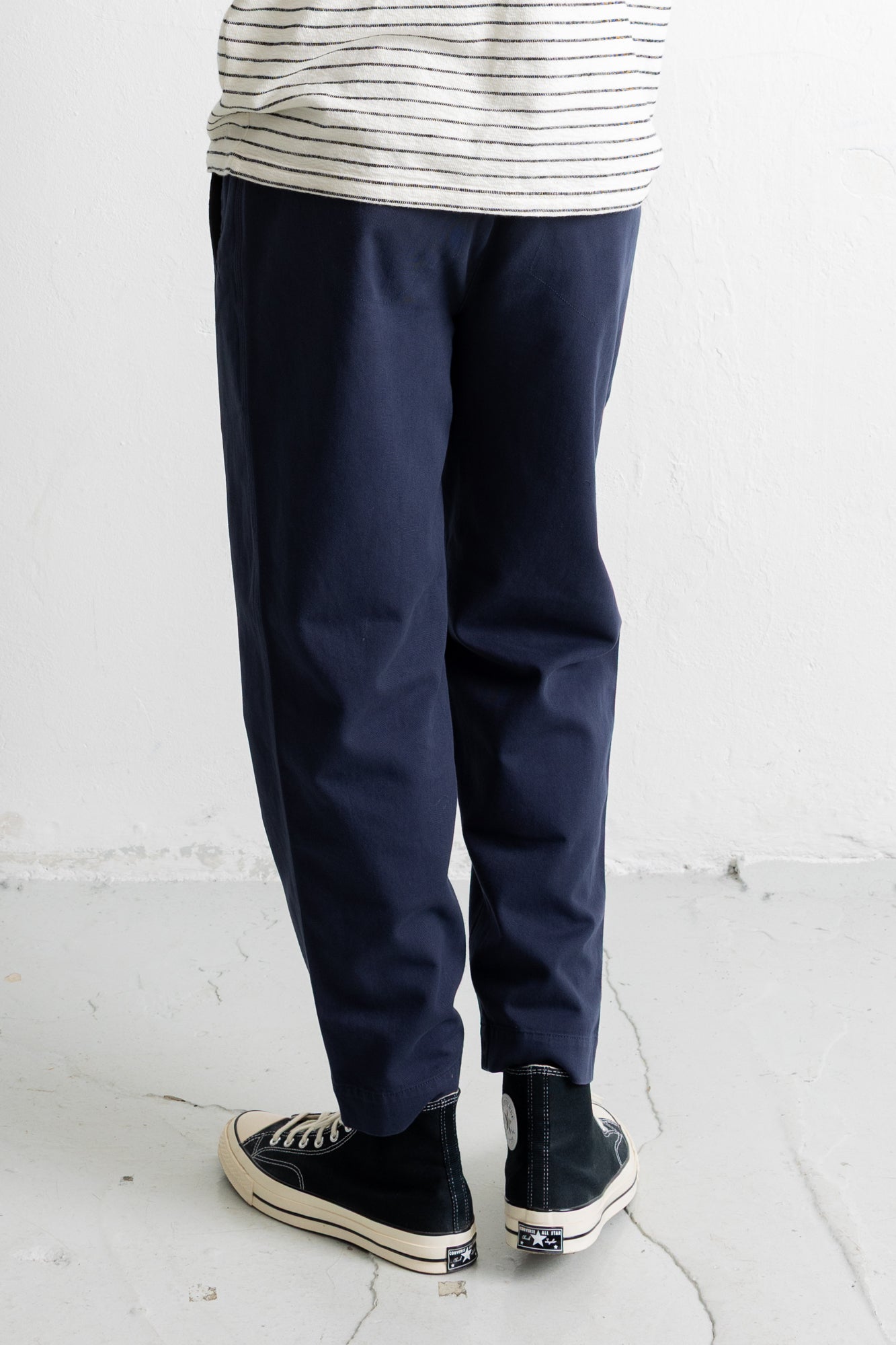A person shown from the waist down wearing navy blue relaxed-fit pants, a cream and navy striped t-shirt, and black high-top sneakers with white soles, standing against a white textured wall.