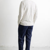 A man viewed from behind wearing a cream long-sleeve shirt with thin dark horizontal stripes, navy blue pants, and black canvas sneakers, standing against a plain white wall.