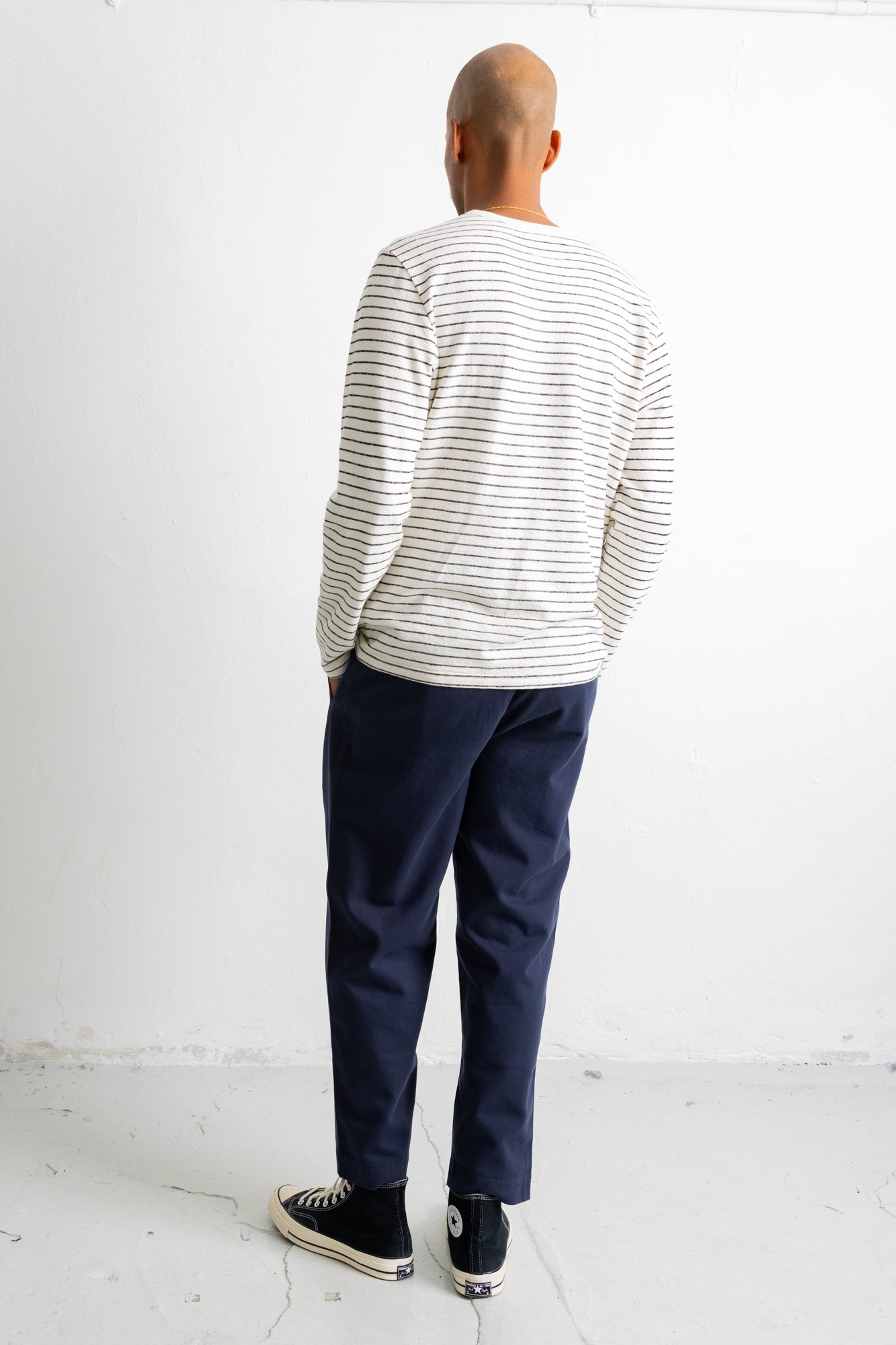 A man viewed from behind wearing a cream long-sleeve shirt with thin dark horizontal stripes, navy blue pants, and black canvas sneakers, standing against a plain white wall.
