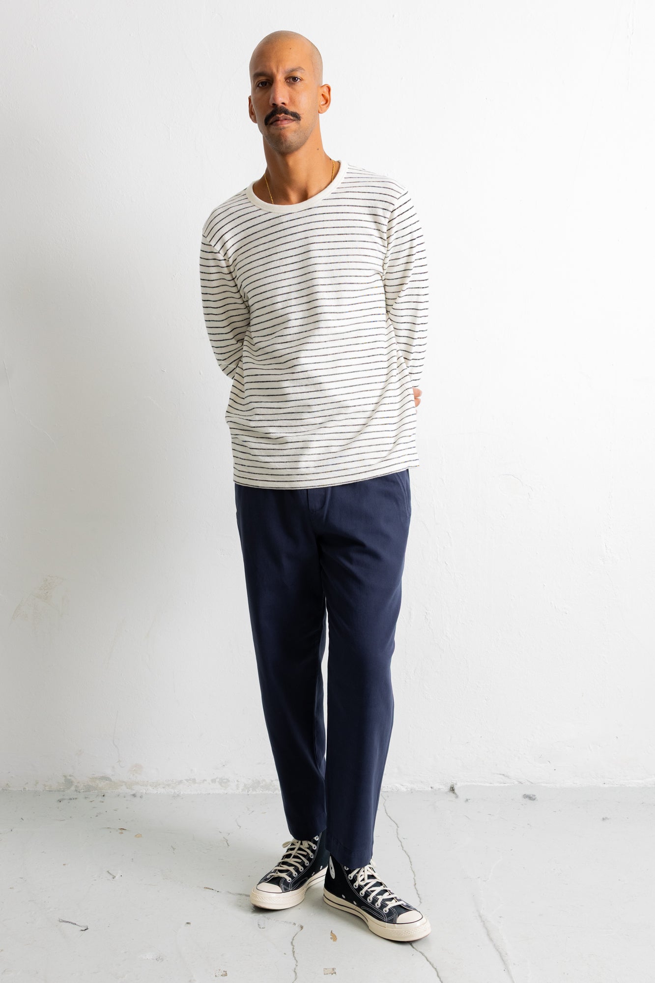 A man wearing a cream and navy striped long-sleeve shirt, navy pants, and black canvas sneakers, standing against a white wall.