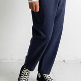 A person wearing navy blue relaxed-fit pants, a white long-sleeve shirt with thin black horizontal stripes, and black low-top canvas sneakers with white soles, standing against a light gray wall.