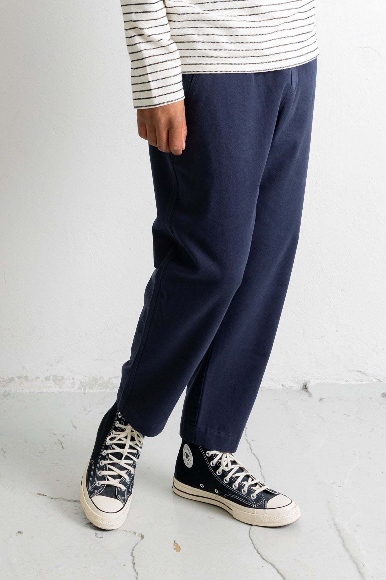 A person wearing navy blue relaxed-fit pants, a white long-sleeve shirt with thin black horizontal stripes, and black low-top canvas sneakers with white soles, standing against a light gray wall.