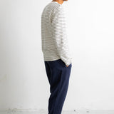 A man in profile wearing a white long-sleeve shirt with thin dark horizontal stripes, navy blue trousers, and black canvas sneakers, standing against a plain white wall.