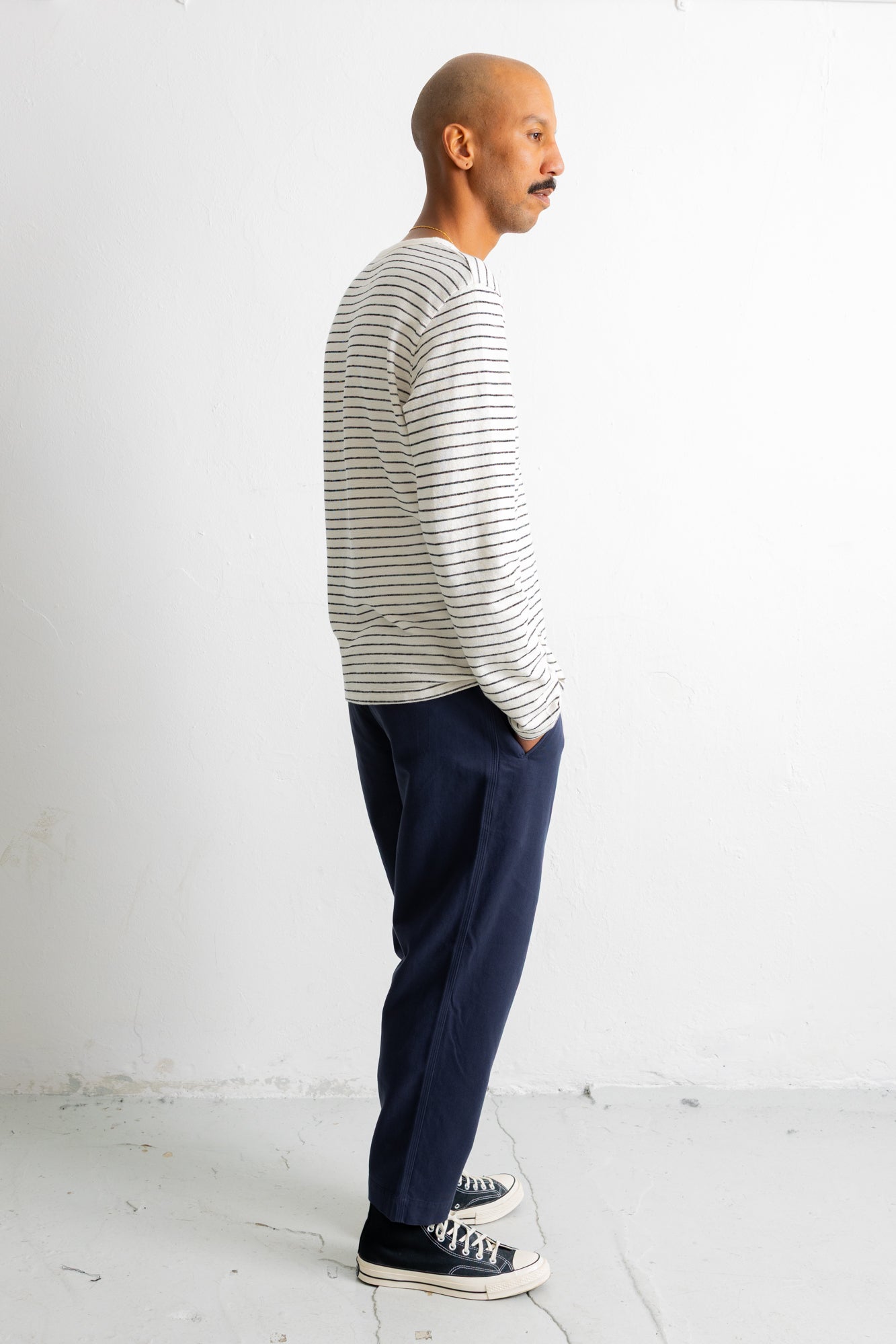 A man in profile wearing a white long-sleeve shirt with thin dark horizontal stripes, navy blue trousers, and black canvas sneakers, standing against a plain white wall.