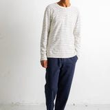 A man wearing a white long-sleeve crewneck shirt with thin navy horizontal stripes, dark navy pants, and black canvas sneakers, standing against a white studio wall.