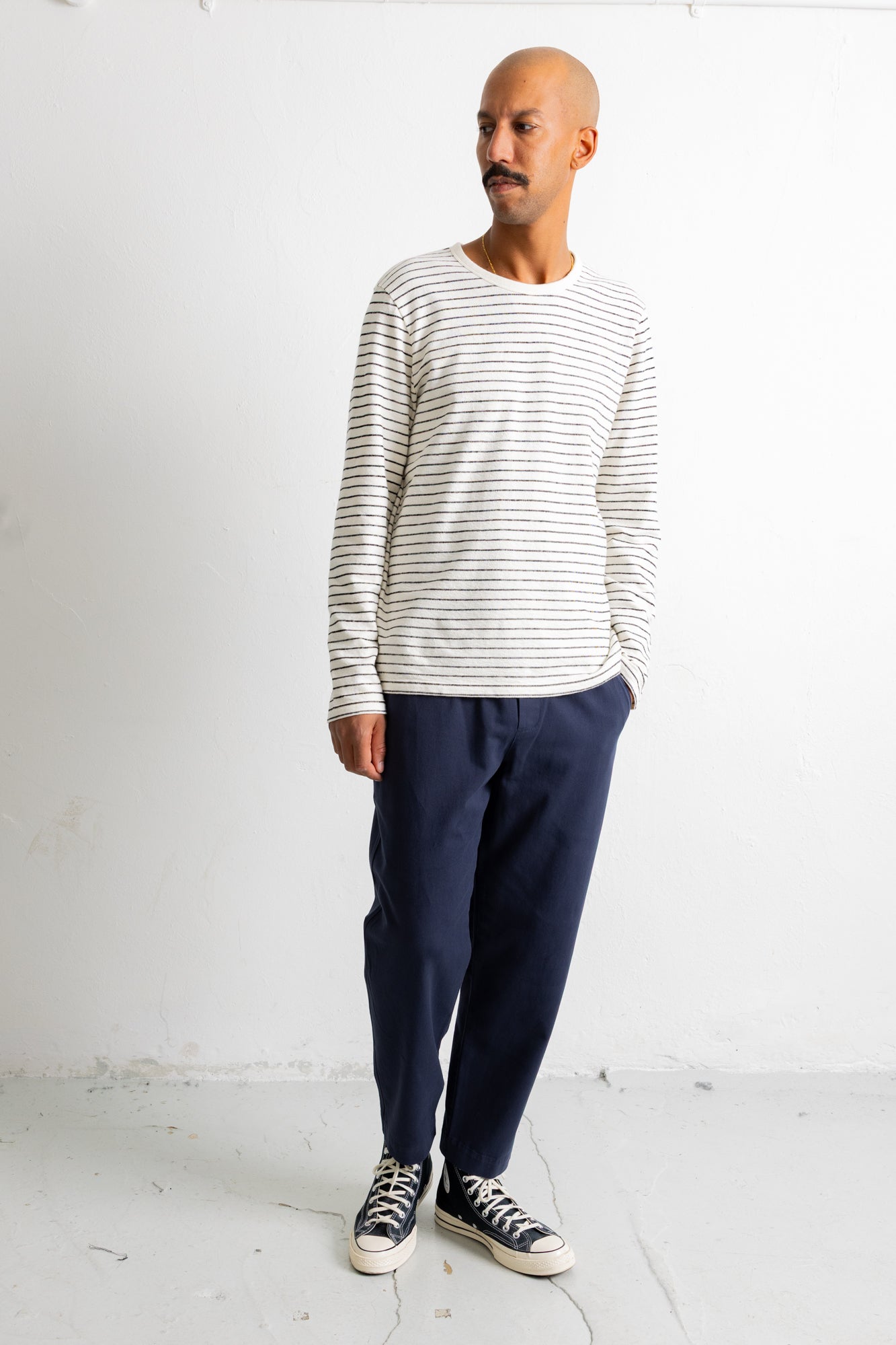 A man wearing a white long-sleeve crewneck shirt with thin navy horizontal stripes, dark navy pants, and black canvas sneakers, standing against a white studio wall.