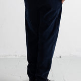 A person shown from the back wearing dark navy corduroy trousers, a dark green jacket, a white undershirt, and black shoes, standing against a white wall.
