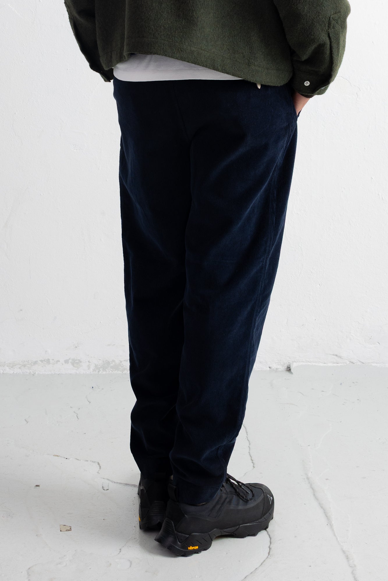 A person shown from the back wearing dark navy corduroy trousers, a dark green jacket, a white undershirt, and black shoes, standing against a white wall.