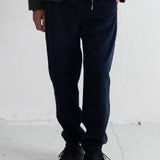 A person wearing dark navy cord pants, a dark olive green jacket, a brown shirt, and black lace-up shoes, standing against a white wall.