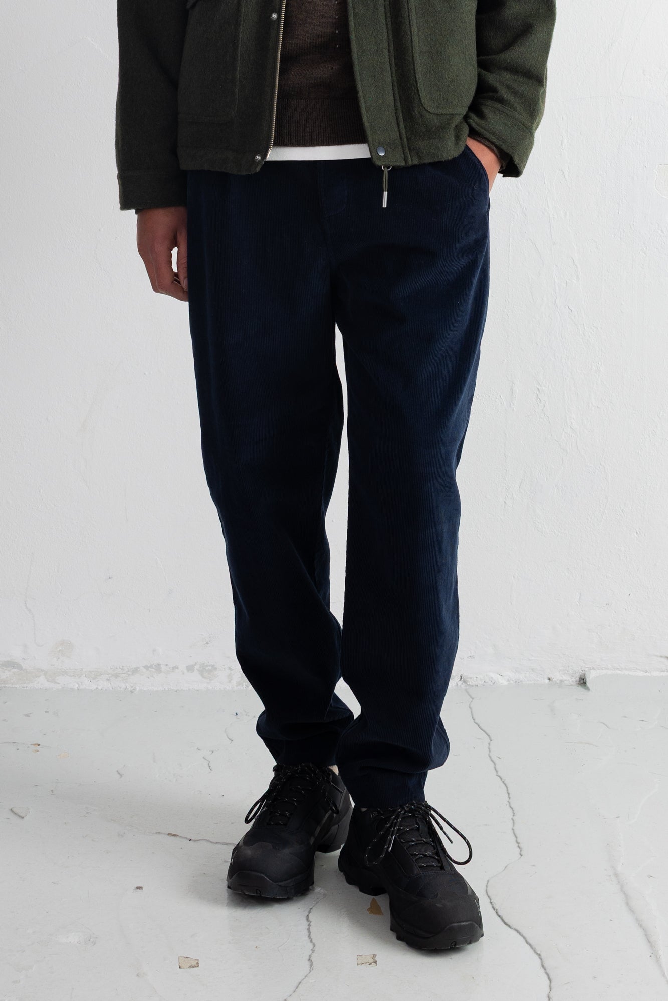 A person wearing dark navy cord pants, a dark olive green jacket, a brown shirt, and black lace-up shoes, standing against a white wall.