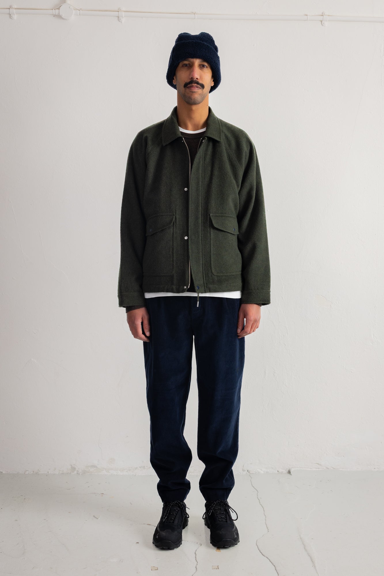 A man stands against a plain white studio wall wearing an olive green utility jacket over a white shirt, dark blue cord trousers, black sneakers, and a dark navy beanie.