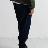A person shown from the chest down wearing dark navy corduroy trousers, a dark olive green jacket, a white undershirt, and black hiking boots against a white wall.