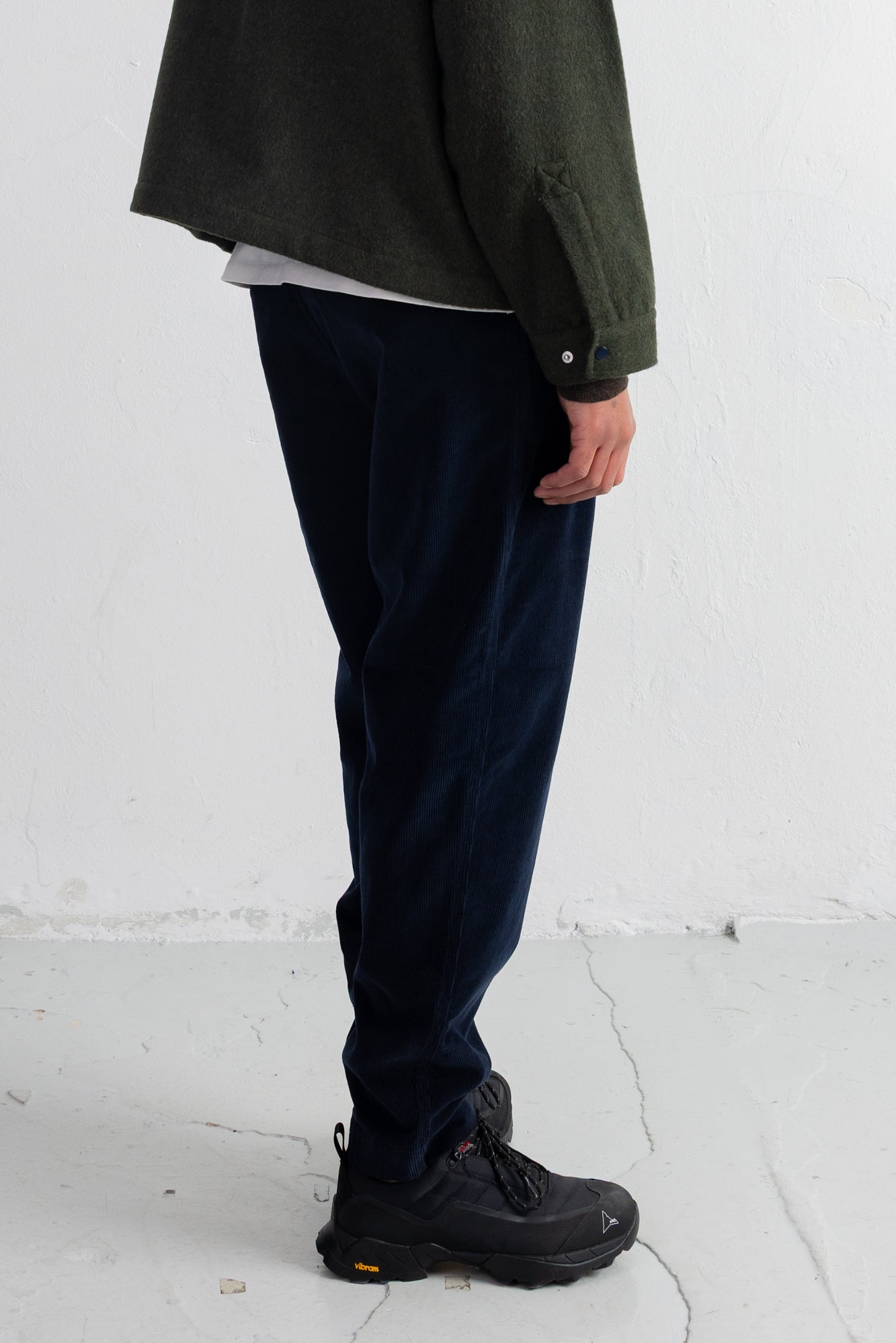 A person shown from the chest down wearing dark navy corduroy trousers, a dark olive green jacket, a white undershirt, and black hiking boots against a white wall.