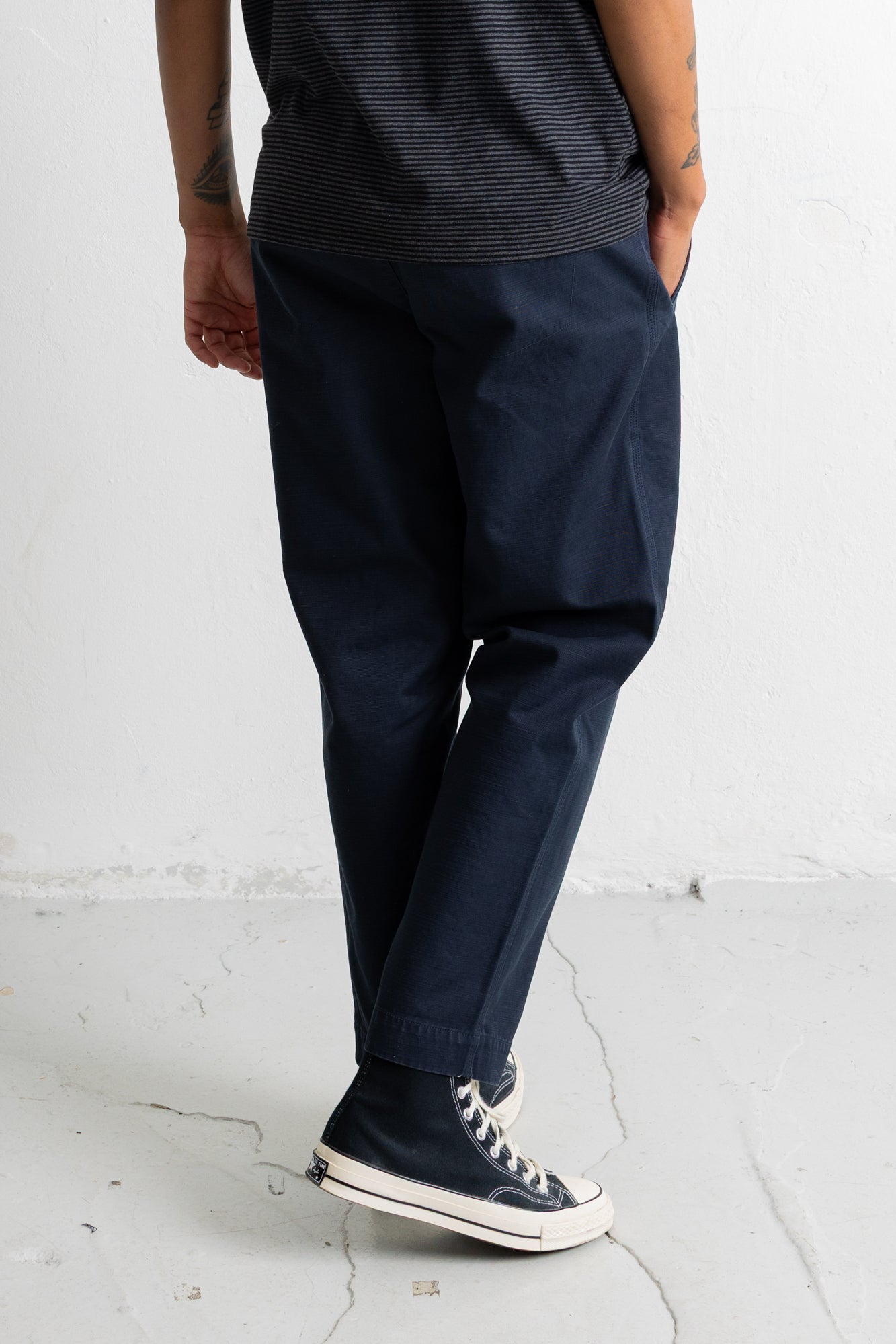 A person wearing dark navy trousers, a dark striped t-shirt, and navy high-top sneakers with white soles, standing against a white wall.