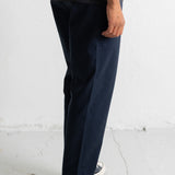 A person wearing dark navy straight-leg trousers, a dark striped t-shirt, and black canvas sneakers, standing against a white wall.