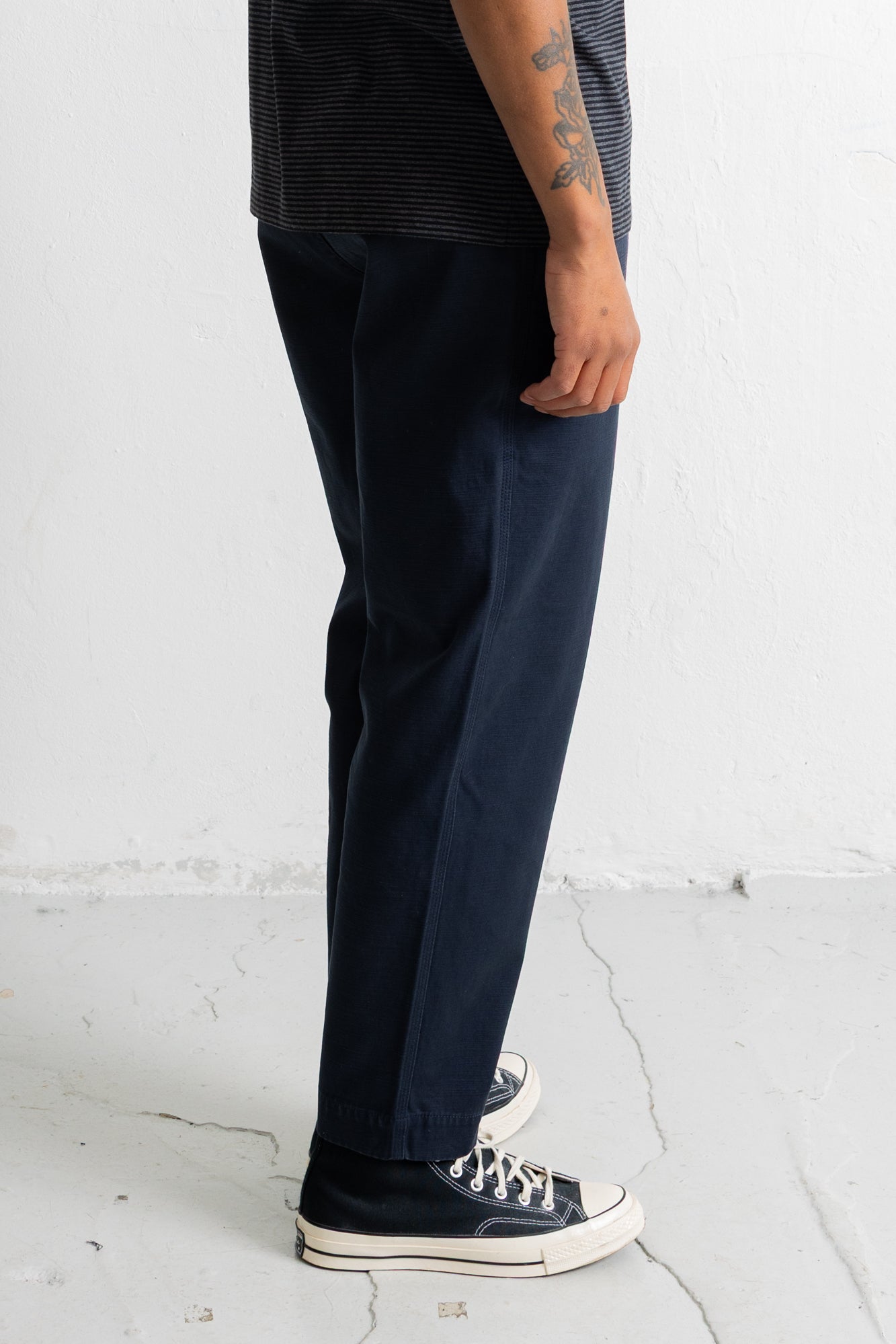 A person wearing dark navy straight-leg trousers, a dark striped t-shirt, and black canvas sneakers, standing against a white wall.