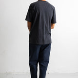 A man viewed from behind wearing a dark charcoal gray textured short-sleeve t-shirt, navy blue pants, and black canvas sneakers, standing against a plain white wall.