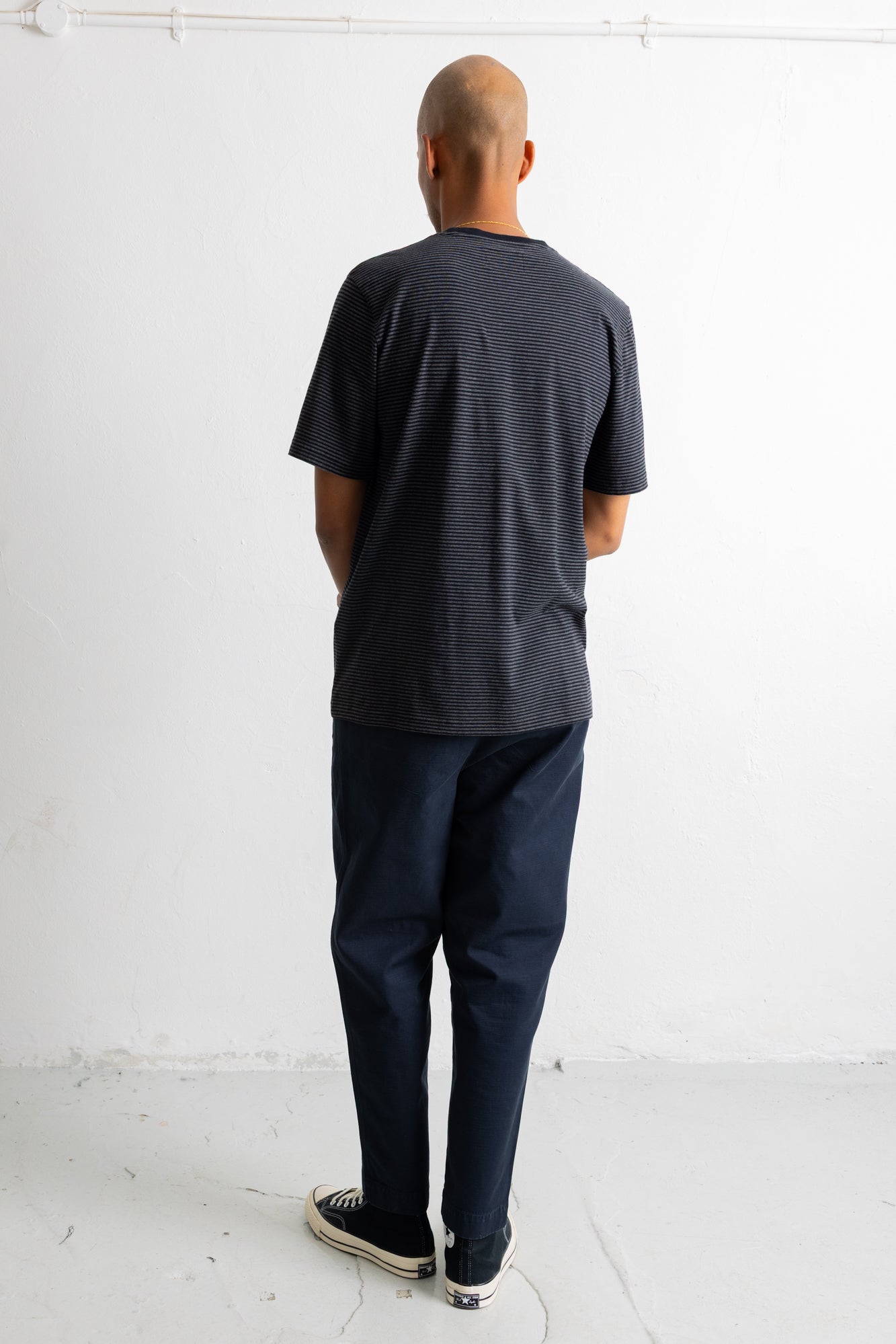 A man viewed from behind wearing a dark charcoal gray textured short-sleeve t-shirt, navy blue pants, and black canvas sneakers, standing against a plain white wall.