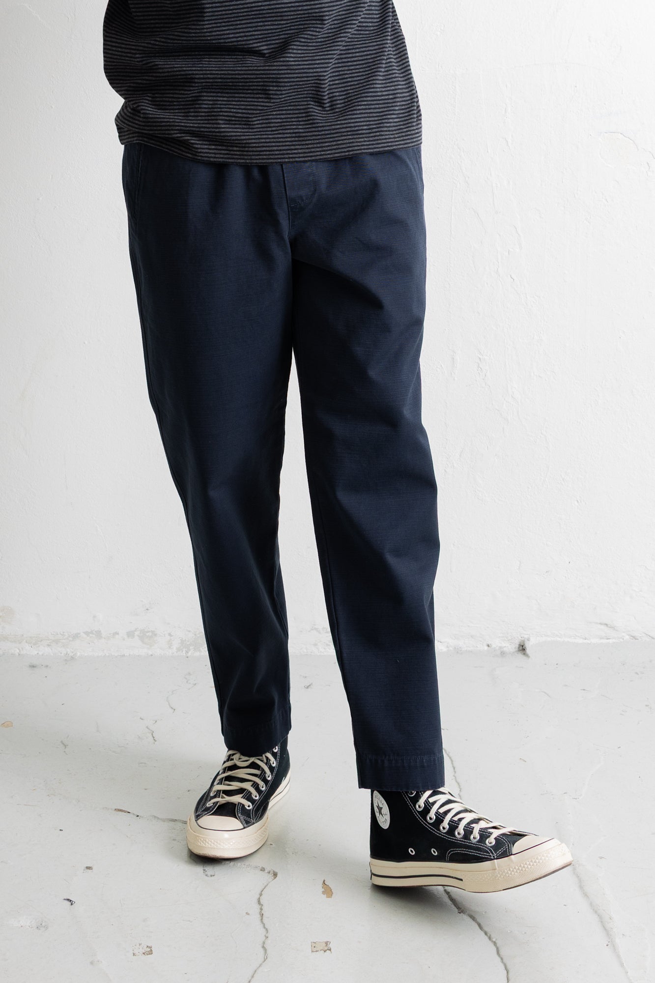 A person wearing dark navy pants, a dark striped shirt, and black canvas sneakers with white soles, standing against a light gray wall.