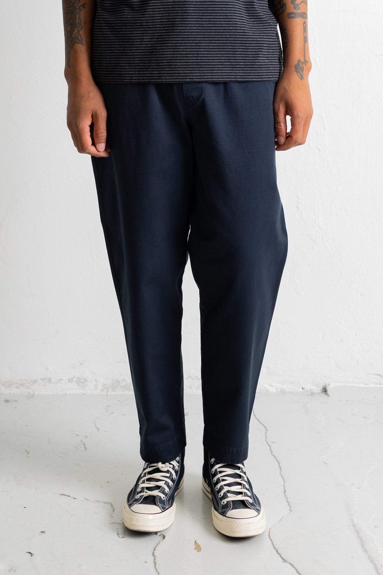 A person wearing dark navy relaxed-fit trousers, a dark striped top, and black canvas sneakers with white soles, standing against a light gray wall.