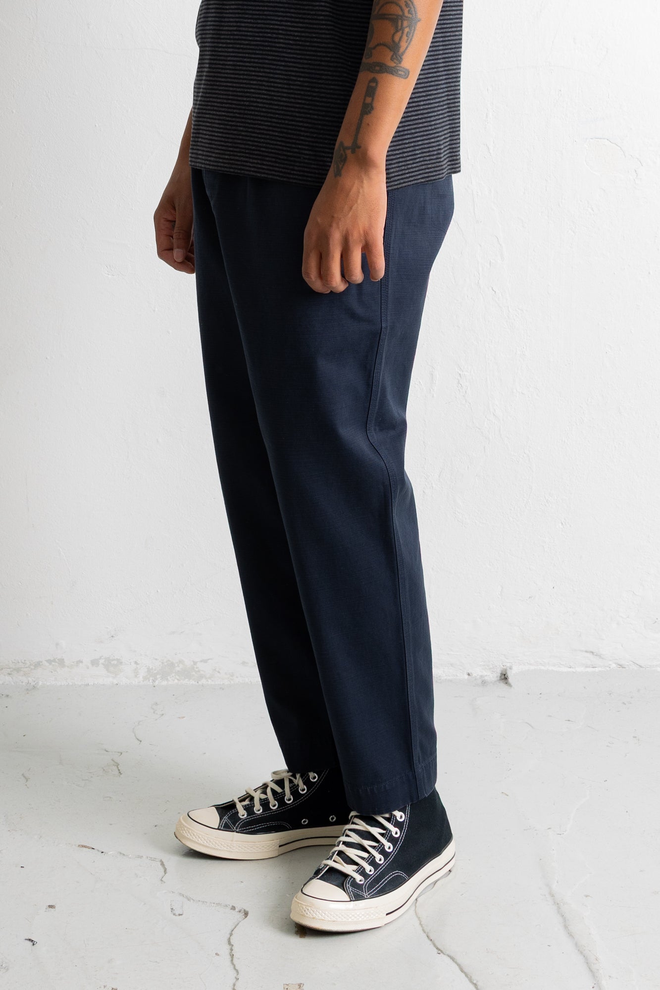 A person wearing dark navy blue relaxed-fit pants, a dark striped t-shirt, and black canvas high-top sneakers, standing against a white wall.