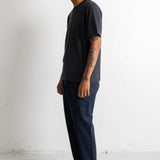 A man in a dark charcoal t-shirt, navy blue pants, and black canvas sneakers stands in profile against a plain white wall.