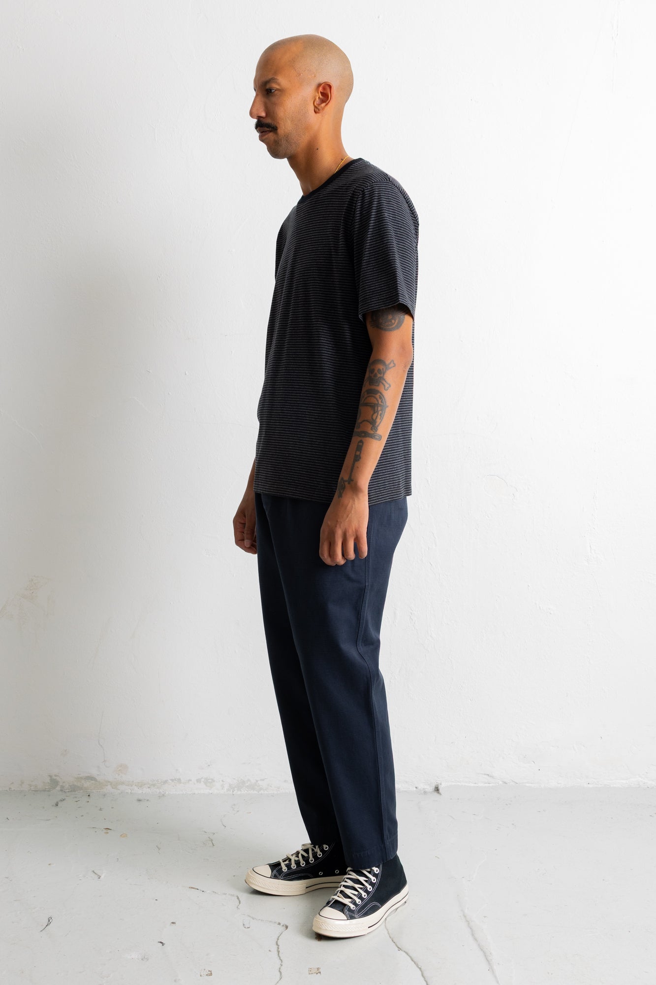 A man in a dark charcoal t-shirt, navy blue pants, and black canvas sneakers stands in profile against a plain white wall.