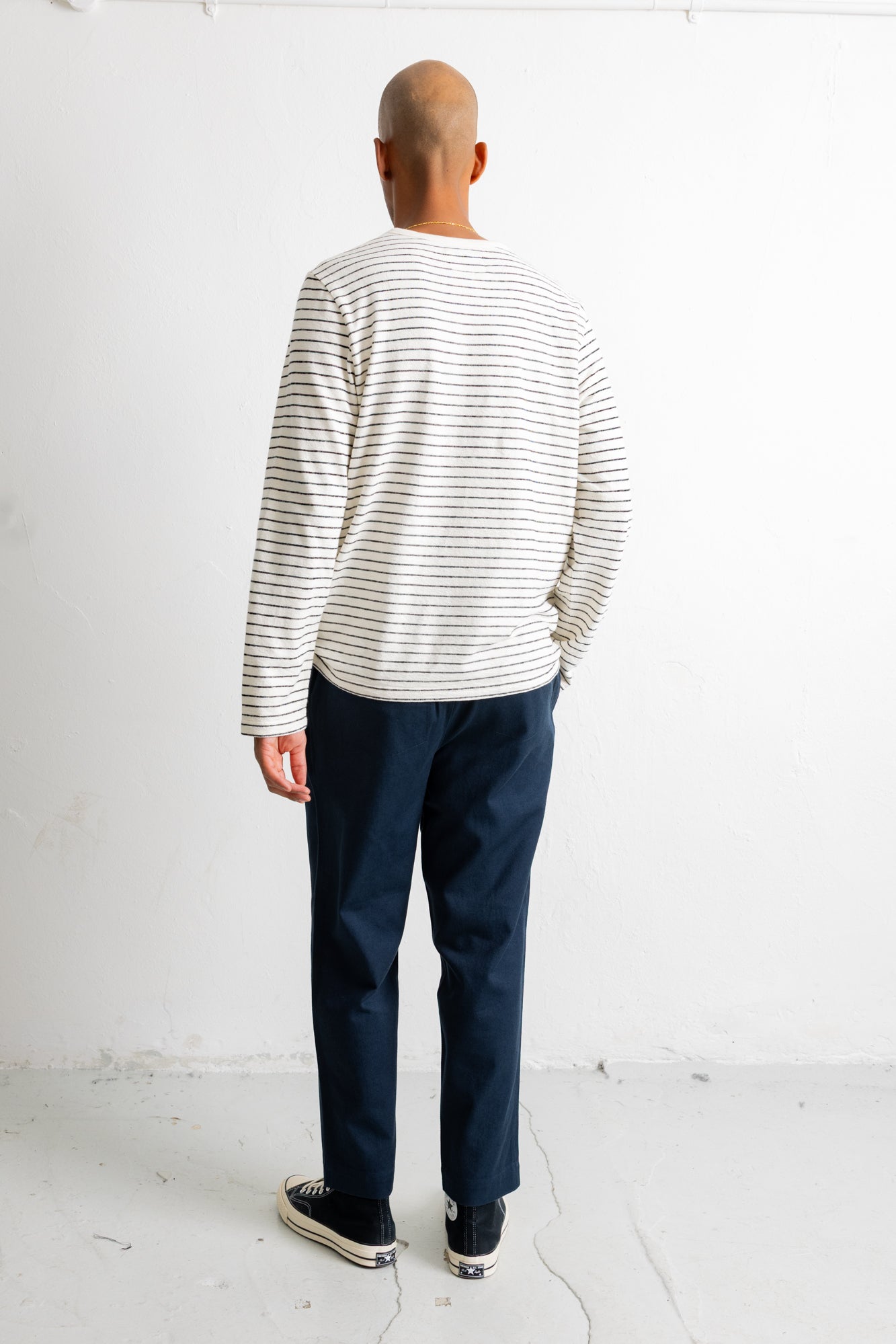 A man viewed from behind wearing a cream long-sleeve shirt with thin dark horizontal stripes, navy blue pants, and black canvas sneakers, standing against a white studio wall.