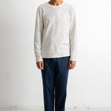 A man stands against a plain white studio wall wearing a cream long-sleeve knit top with thin dark horizontal stripes, dark navy trousers, and black-and-white sneakers.