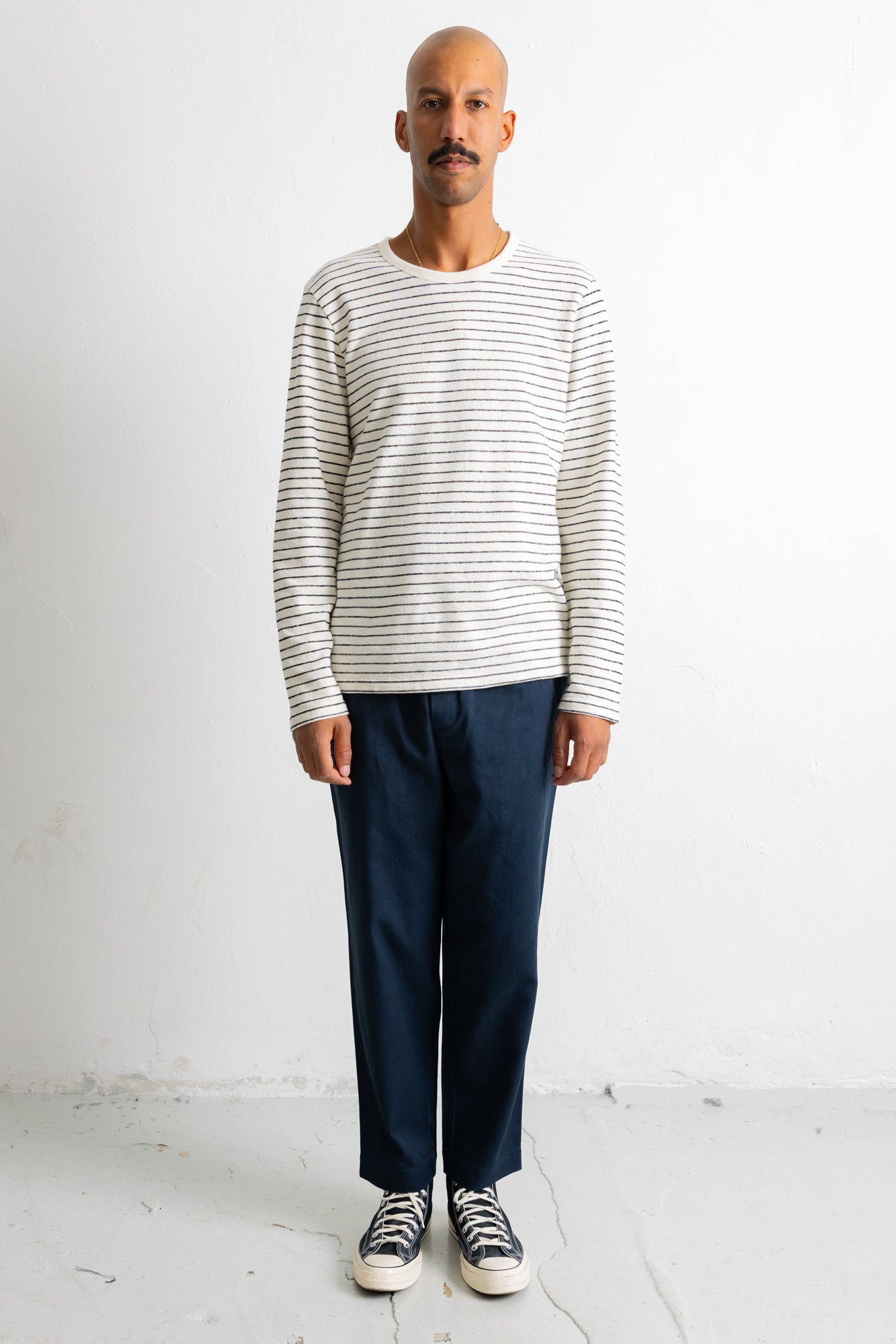 A man stands against a plain white studio wall wearing a cream long-sleeve knit top with thin dark horizontal stripes, dark navy trousers, and black-and-white sneakers.