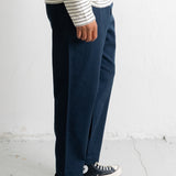A person shown from the waist down wearing dark navy wide-leg trousers, a white long-sleeve shirt with thin black horizontal stripes, and black low-top sneakers with white soles, standing against a white wall.