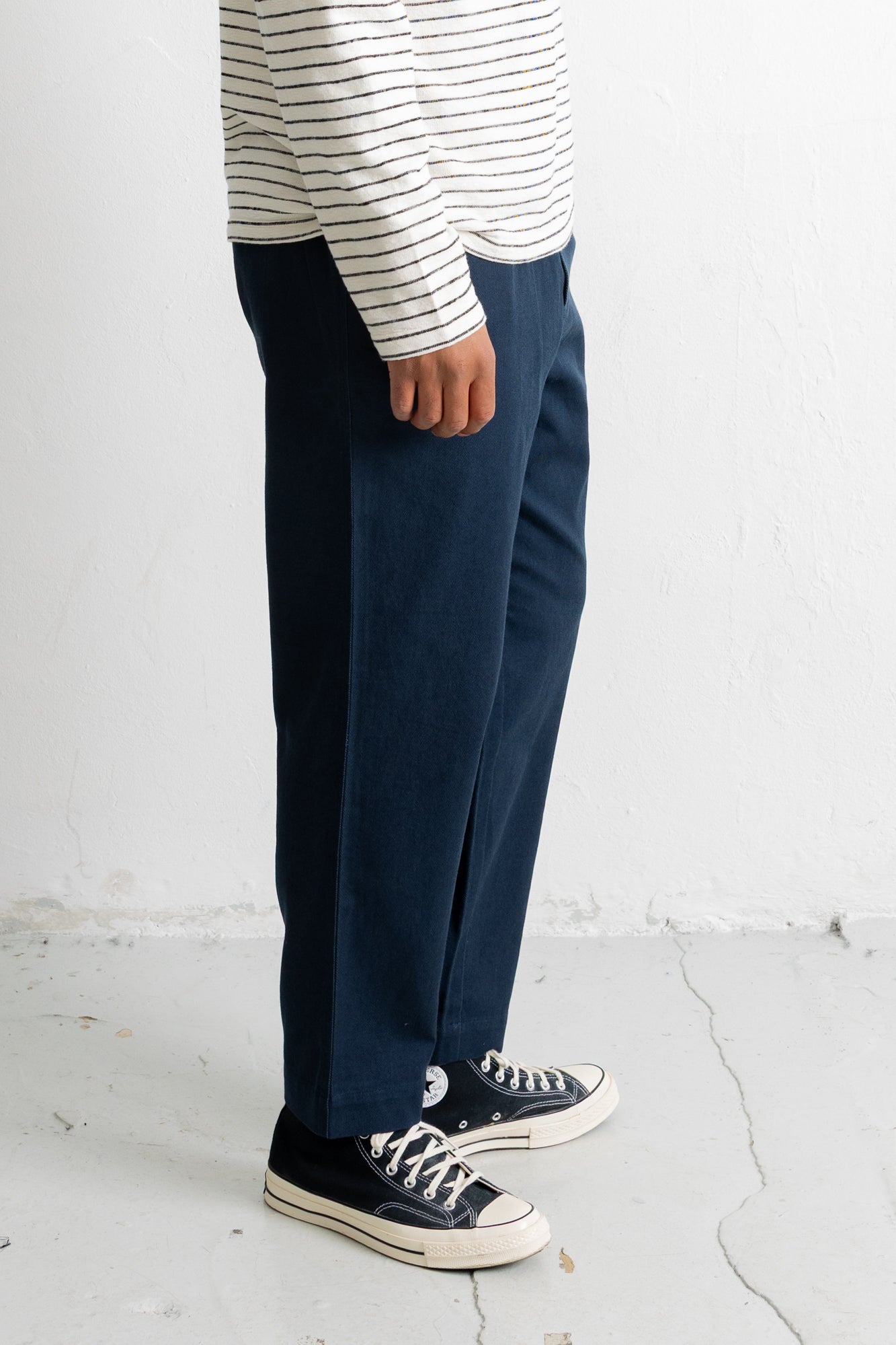 A person shown from the waist down wearing dark navy wide-leg trousers, a white long-sleeve shirt with thin black horizontal stripes, and black low-top sneakers with white soles, standing against a white wall.