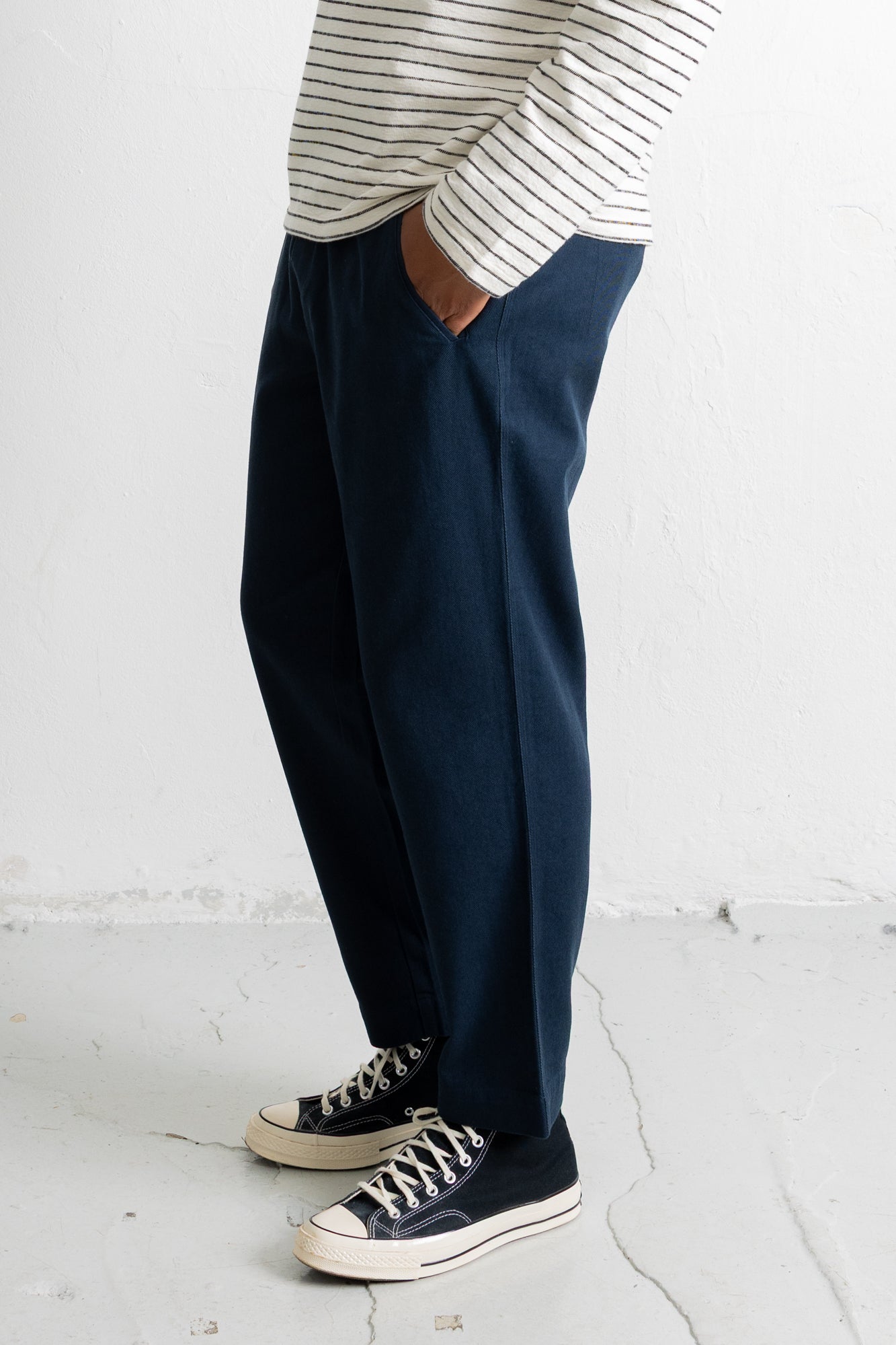 A person wearing dark navy wide-leg trousers, a cream and navy striped long-sleeve shirt, and navy canvas sneakers with white rubber soles, standing against a white wall.